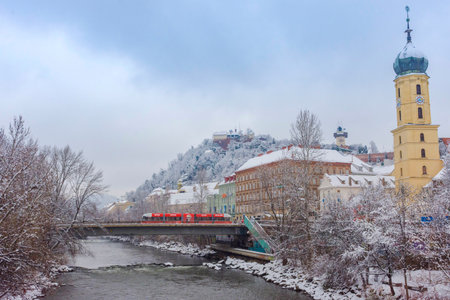 Graz, Austria-December 09, 2021: Mur river, the Franciscan Church tower and the famous clock tower in the background, in the city center of Graz, Steiermark, Austria, with snow, in winterのeditorial素材