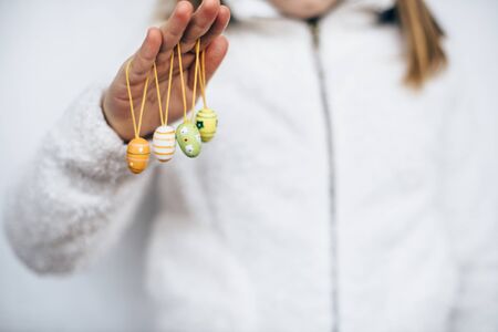 Girl in white zipped-up jumper with Easter egg decorations hanging from fingers of hands raisedの写真素材