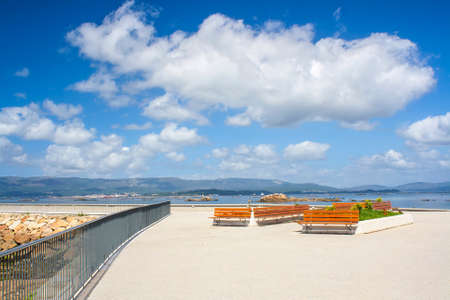 Benches on the boardwalk of Vilanova de Arousa with white cumulus cloud on the blue skyの写真素材
