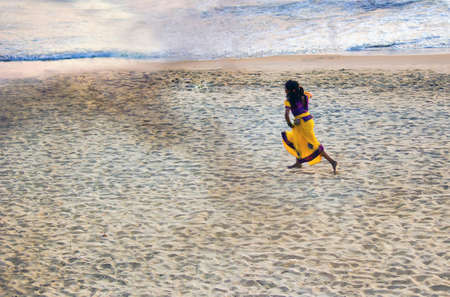 A small girl in a traditional Indian yellow dress runs cheerfully in sandy beach in Kerala, located in South Indiaのeditorial素材