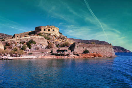 Crete, Greece: Wide angle view of Spinalonga unhabited island with a 16th century venetian fortress and the ruins of a formar leper colony against cloudy blue sky and blue water in the foregroundのeditorial素材