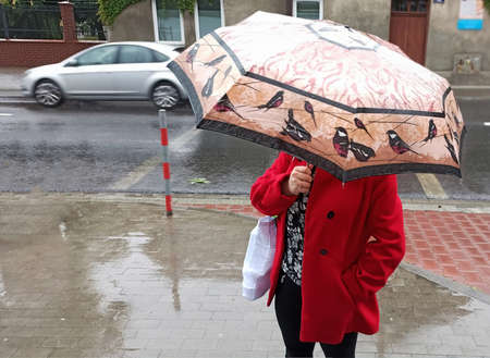 A lady or female in red coat with umbrella standing in the rain during winter in European city with car moving on road in the backgroundの写真素材