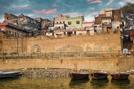 Varanasi, India - November 01, 2016: Bunch of wooden boats docked on river Ganges against a wall with graffiti art and old building exterior architecture during dramatic cloud and sunset or sunriseのeditorial素材