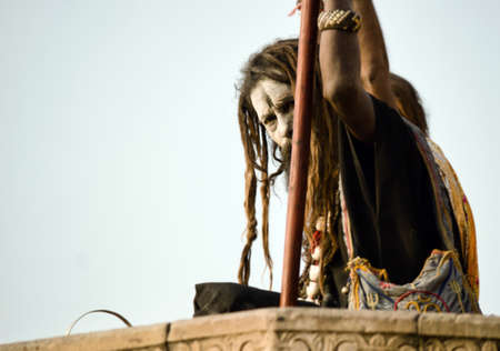 Varanasi, India - November 01, 2016: A hindu bearded sadhu, pilgrim or Aghori baba looking straight sitting with a stick against ganges river during day with dreadlocksのeditorial素材