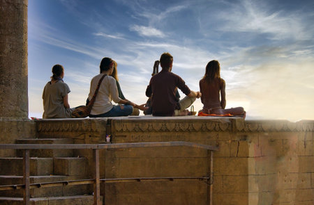 Varanasi, India - November 01, 2016: Bunch of tourists, sadhu baba and pilgrims in sitting posture meditating together on a pile of rock during morning facing ganges river and sunrise. Uttar pradeshのeditorial素材