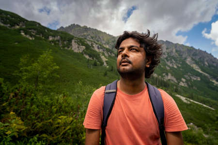 Portrait of an adventurer Indian tourist man looking up towards the way forward with bag pack against mountains during hike. Summer adventure and sports conceptの写真素材