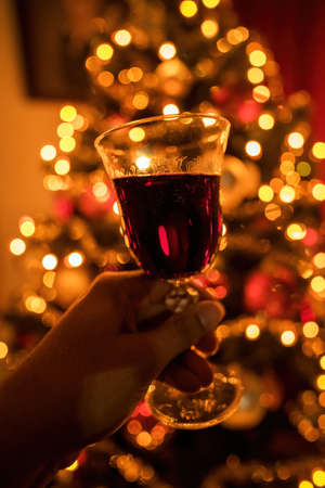 Hand cheering with red wine glass against out of focus christmas tree lights, xmas festive celebration concept.の写真素材