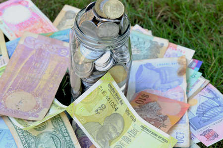 glass jar full of coins on different currency note on green grass background