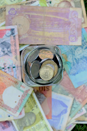 glass jar full of coins on different currency note on green grass background
