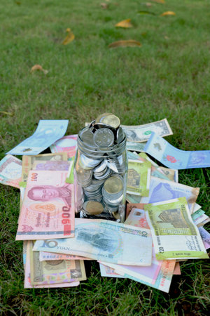 glass jar full of coins on different currency note on green grass background