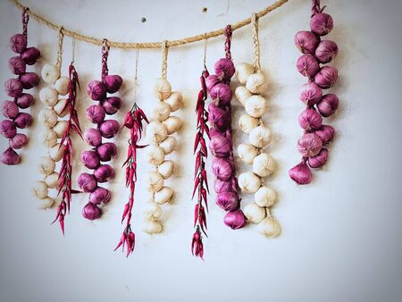 Red Onions, Garlic and Chili necklaces hanging on the rope, isolated in white concrete background. Decoration in Thai restaurant. Ingredient for cooking Thai spicy food. Thai food concept.の写真素材
