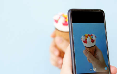 A smartphone take a photo of Cone ice cream with multicolored candy on topping carry on hand isolated in blue background, Selective focus blurred background has copy space for food business concept.の写真素材