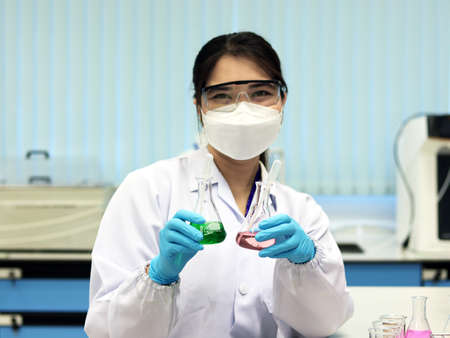 The scientist wear PPE and holds an Erlenmeyer flask laboratory, with green and pale pink solvent solution from reaction indicator acid titration experiment, analysis compounding in wastewater sample.の写真素材