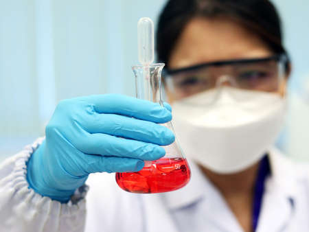 The scientist wears PPE and holds an Erlenmeyer flask laboratory, with red solvent solution from reaction methyl orange indicator acid titration experiment, analysis compounding in wastewater sample.の写真素材