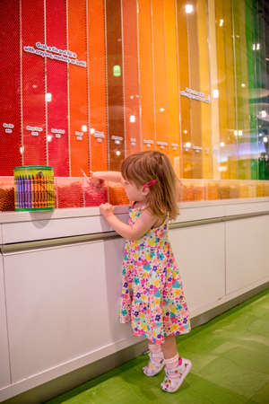 Little girl standing on tiptoe in a colorful art supply store selecting colored wax crayons from a display with wall stripes in an extensive range of colors and hues in the spectrumのeditorial素材