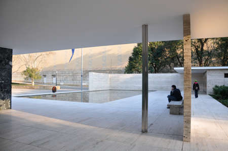 Reflecting pool in geometric building with man seated on stone bench nearby as woman walks towards exit in Barcelona Pavilion by Ludwig Mies van der Roheのeditorial素材