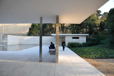 Man contemplates as he sits in patio by reflecting pool as woman leaves via corridor behind him in Barcelona Pavilion by Ludwig Mies van der Roheのeditorial素材