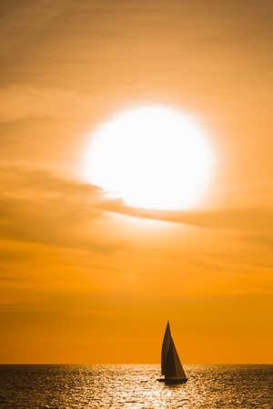 Sailboat sailing on the ocean during a spectacular orange tropical sunset silhouetted against the vivid sky in a reflected path of sunlight on the waterの写真素材