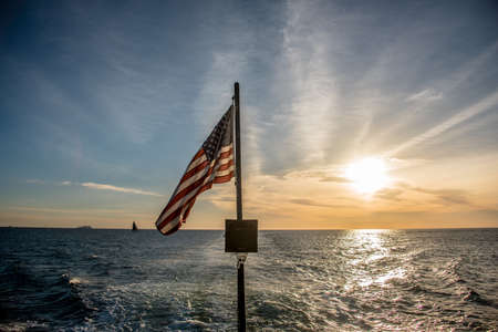 American flag flying from a sailboat at sea viewed over the stern against a sunset sky with a wake in the water behind the boatの写真素材