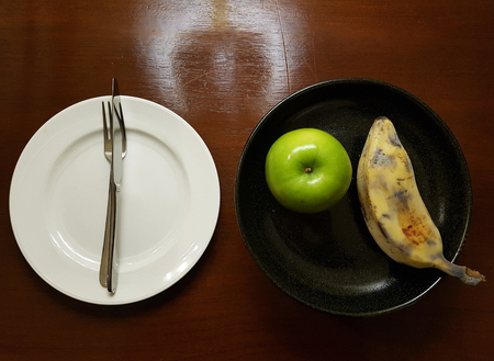 Fresh fruit (banana and green apple) on the black plate and white plate with fork and knife - on the wooden table with a little light reflect from the lampの写真素材