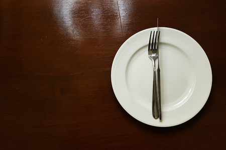 White ceramic plate with fork and knife on the wooden table - a little light reflect from lampの写真素材