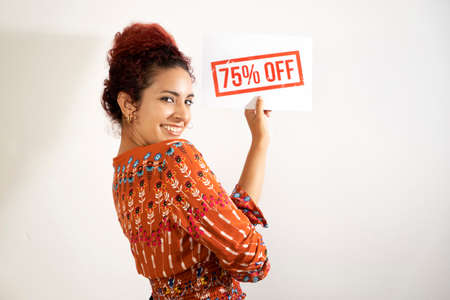 Excited girl smiling and holding a 75% discount sign with one hand. Young woman with white skin and curly red hair dressed in colorful patterned clothingの写真素材