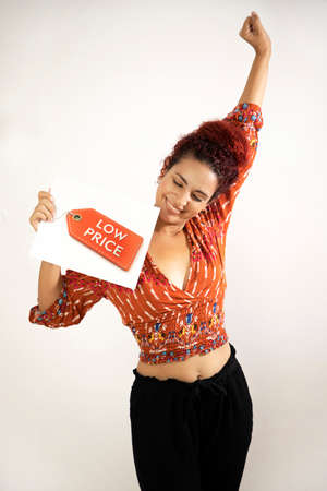 Overjoyed girl smiling, raising one hand with a clenched fist and holding a low price sign with her...