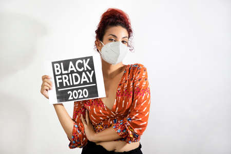 Girl with mask holding a black friday 2020 poster with her hand. Young woman with white skin and curly red hair dressed in colorful patterned clothingの写真素材
