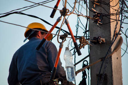 Electric lineman in a yellow helmet working with clamps at heights on the cables of the streets,...