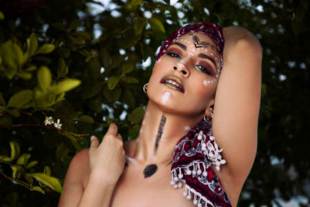 Girl with her face painted as a native inhabitant of Africa or the Amazon, with a scarf on her head, among vegetation, beautiful aboriginalの写真素材