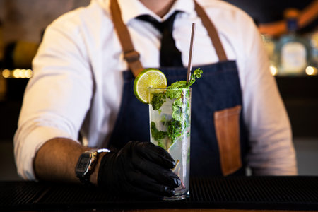 Detail of the hands of a bartender with black gloves placing a crystal glass with a cocktail inside,...
