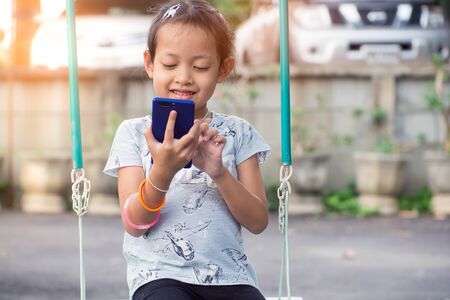 Asian child little girl playing smartphone with happy on the swing.の写真素材