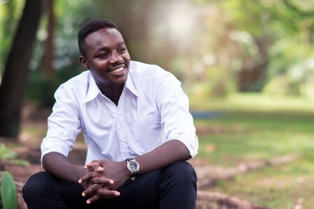 African business man in white shirt smiling and sitting outside with green trees on background.の写真素材