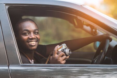 African man driver holding a film camera and smiling while sitting in a car with open front window.の写真素材