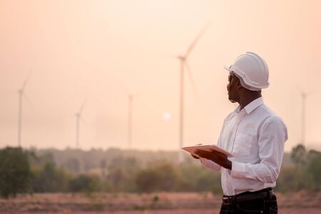 African engineer wearing white hard hat standing with digital tablet against wind turbineの写真素材