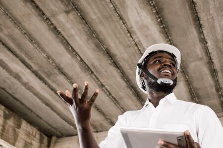 African construction engineer talking and holding tablet while wearing helmet.
