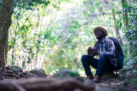 African freedom man traveler holding camera with backpack sitting in the green natural forest.のeditorial素材