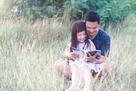 Asian father sitting on grass with little daughter sitting on him and reading a book.の写真素材