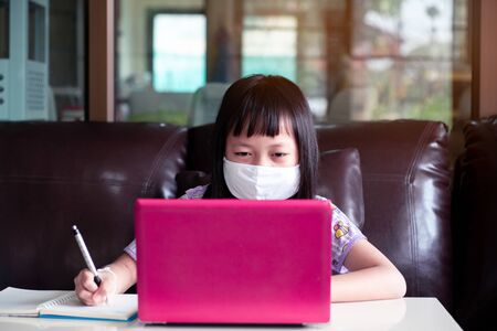 Asian child girl studying homework and wearing face mask during her online lesson at home for protect 2019-nCoV or Covid 19 virus , online education concept.の写真素材