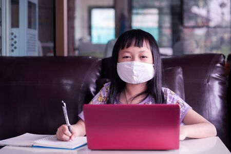 Asian child girl studying homework and wearing face mask during her online lesson at home for protect 2019-nCoV or Covid 19 virus , online education concept.の写真素材