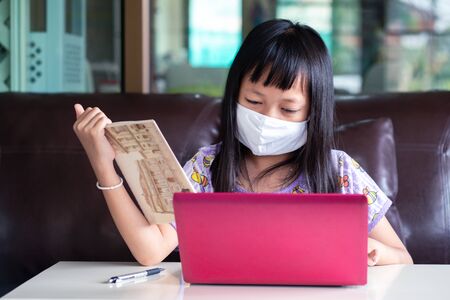 Asian child girl studying homework and wearing face mask during her online lesson at home for protect 2019-nCoV or Covid 19 virus , online education concept.の写真素材