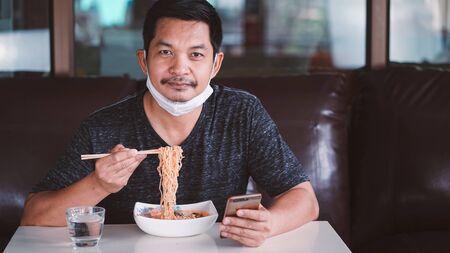 Man in face mask eating a noodle and holding a smart phone at home . Stay at home for protect 2019-ncov virus or Covid-19 Concept.の写真素材