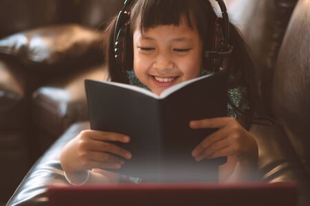 Happy little child girl lying on sofa with learning by  laptop and writing a book at home,Social...