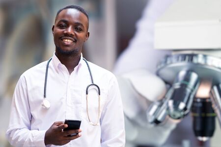 African doctor  holding a smartphone and stethoscope with microscope lab in backgroundの写真素材