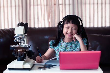 Asian little child  girl wearing headphones learning online by using laptop and microscope at home ,  Online education concept, Social distance during quarantineの写真素材