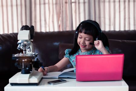 Asian little child  girl wearing headphones learning online by using laptop and microscope at home ,  Online education concept, Social distance during quarantineの写真素材