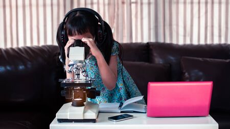 Asian little child  girl wearing headphones learning online by using laptop and looking microscope at home ,  Online education concept, Social distance during quarantineの写真素材