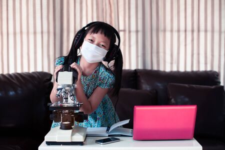 Asian little child  girl wearing headphones learning online by using laptop and looking microscope at home ,  Online education concept, Social distance during quarantineの写真素材