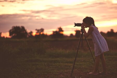 Asian little girl photographer taking picture silhouette sunset sky by digital camera on tripod standing with happy.の写真素材