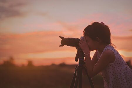 Asian little girl photographer taking picture silhouette sunset sky by digital camera on tripod...